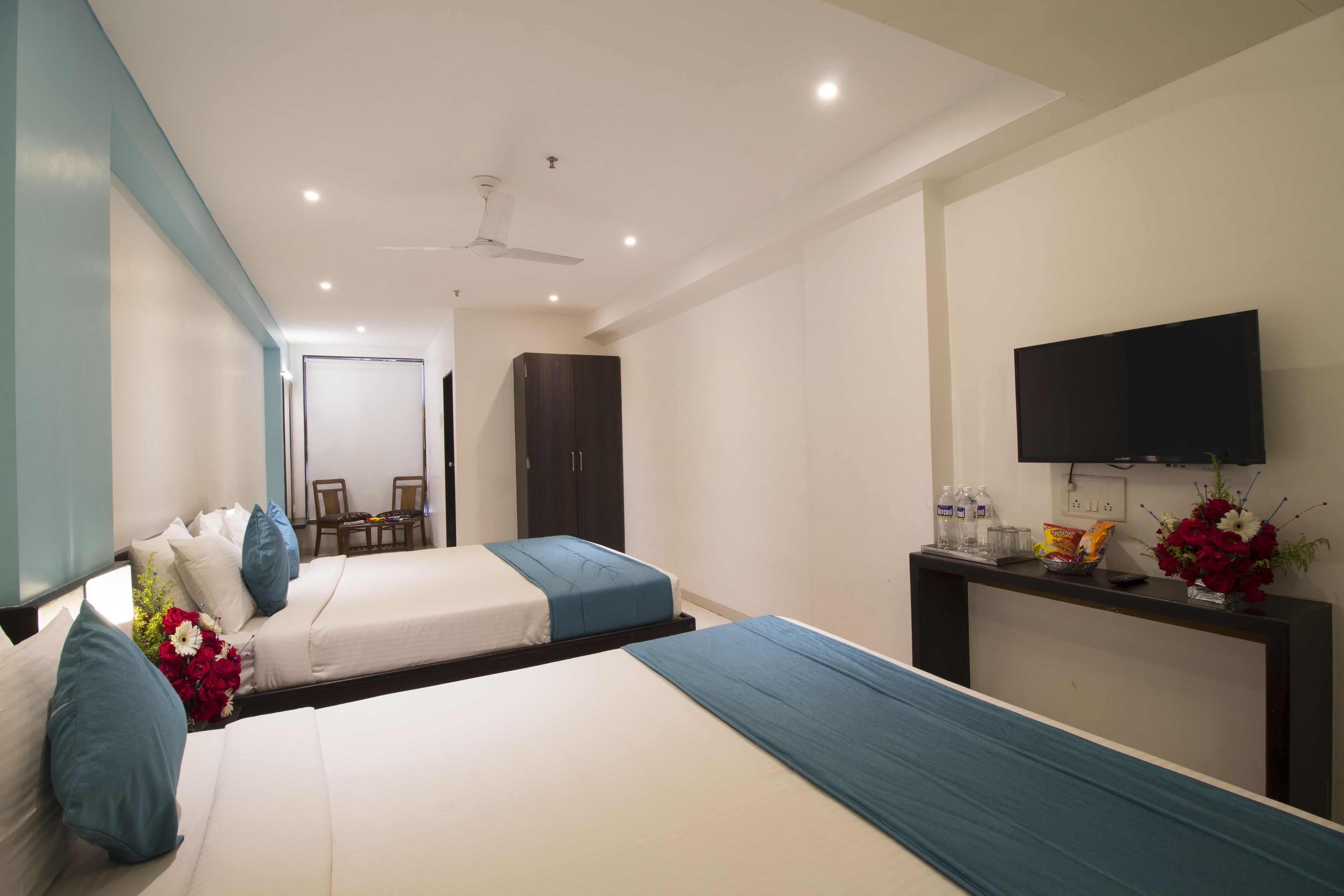 A hotel room with two beds, a TV, a desk with a vase of flowers, and a ceiling fan.