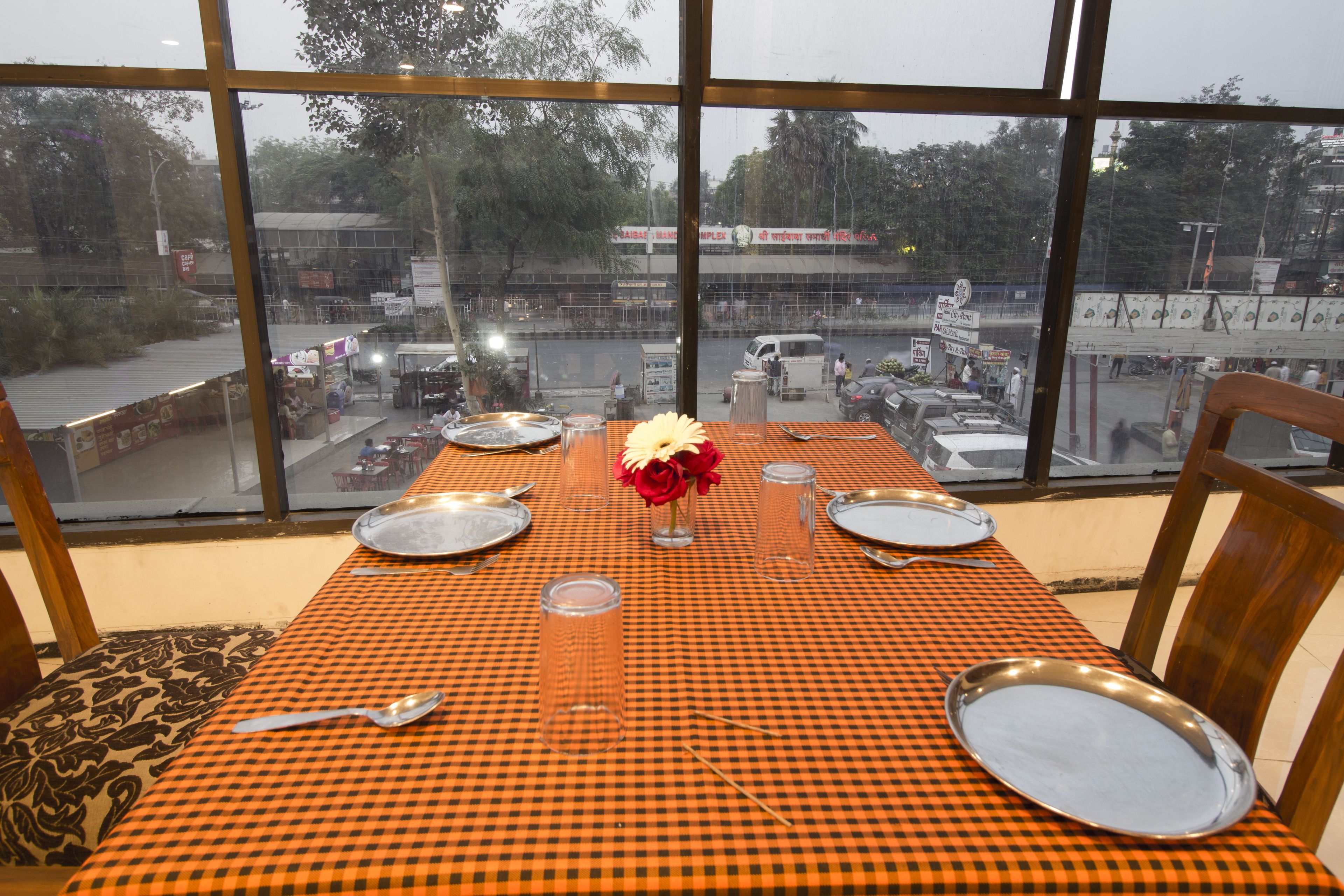A table set for two with a checkered tablecloth, a vase with flowers, and a view of a busy street through large windows.