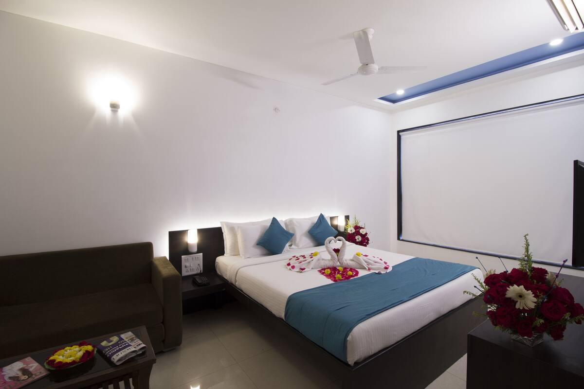 A modern hotel room with a neatly made bed, a flat-screen TV, a ceiling fan, and a small table with a magazine and a bowl of fruit.