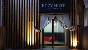Das Rozy Hotel in Namba: Eine moderne Fassade mit vertikalen Holzlamellen und ein markanter Eingangsbereich mit Glastür und weißen Vorhängen.