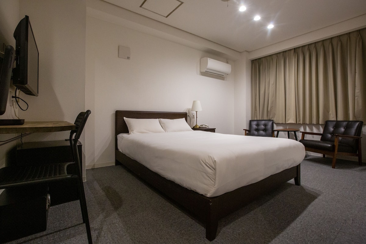 A hotel room with a large bed, a desk with a chair, a TV, a lamp, and a window with curtains.