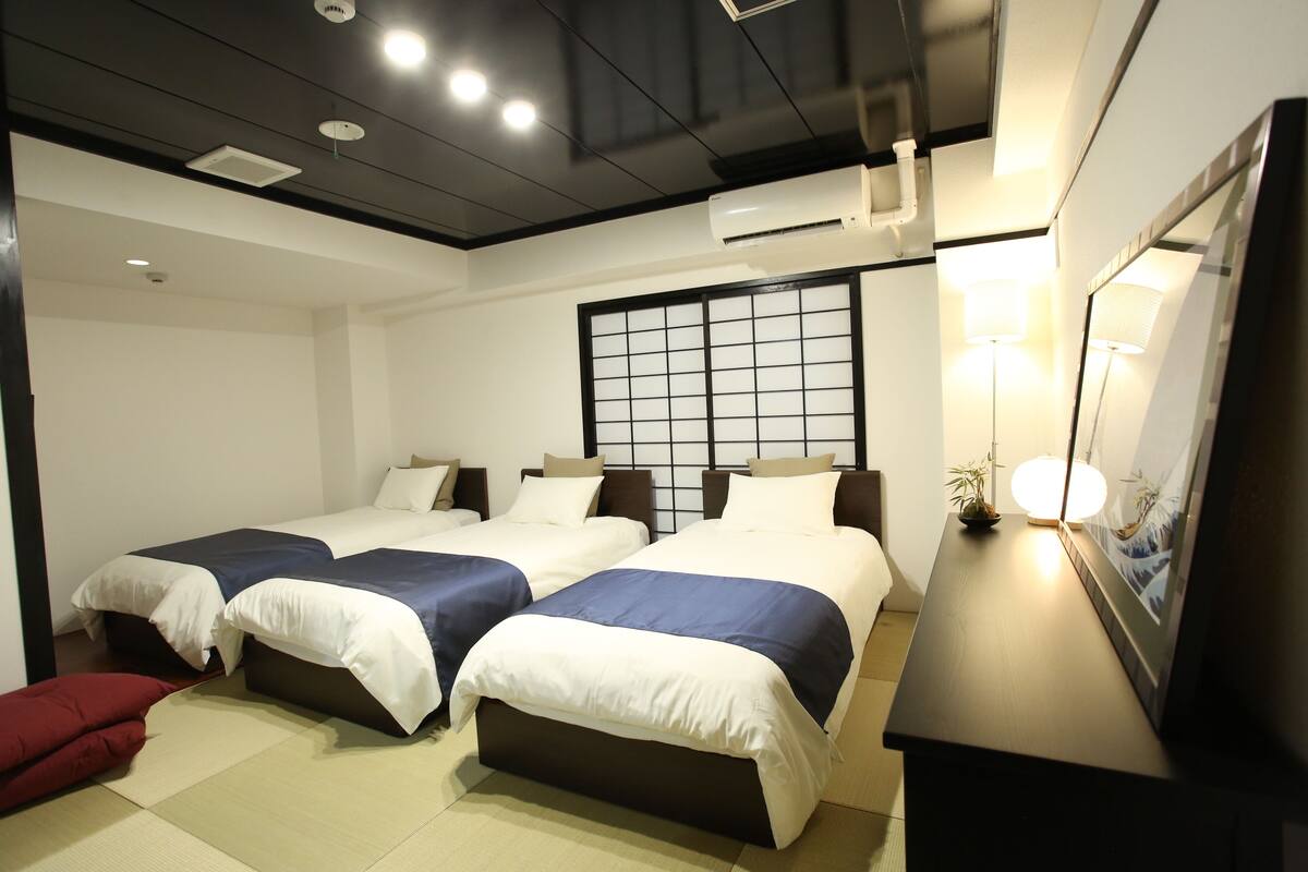 A hotel room with two beds, a large window with a grid pattern, a bedside table with a lamp, and a TV on a stand.