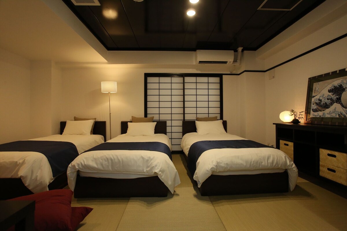 Et hotelværelse med tre senge, en traditionel japansk shoji-skærm, et skrivebord med skuffer og en vægmonteret aircondition.