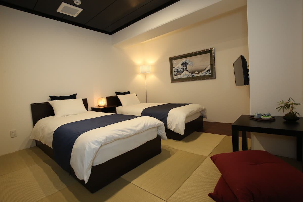 A hotel room with two beds, a bedside table, a wall-mounted TV, a painting, and a small table with a plant.