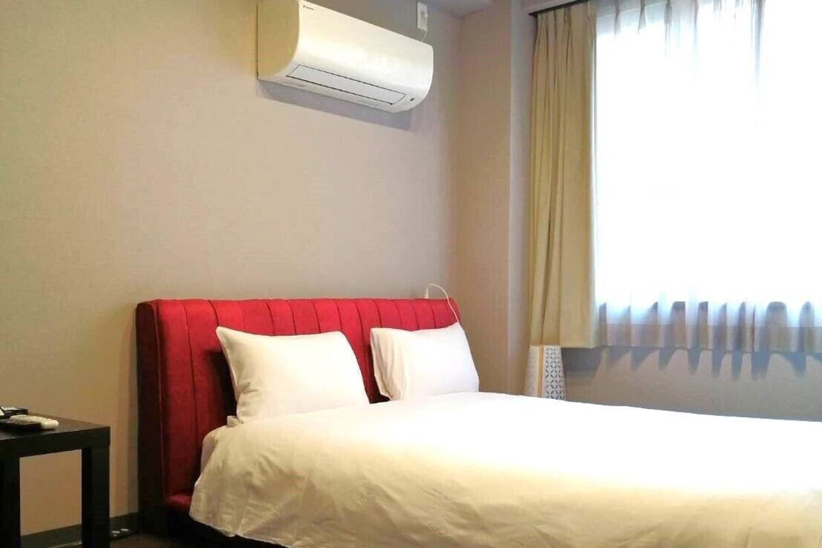 A hotel room with a single bed, a red headboard, a white mattress, a small table, and a window with curtains.