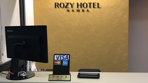 Eine Rezeption im Rozy Hotel Namba mit einem Lenovo-Monitor, einem Schild, das akzeptierte Zahlungsmethoden (VISA, MasterCard, American Express) anzeigt, und einem Schild in japanischer Schrift, das 'Empfang' bedeutet.