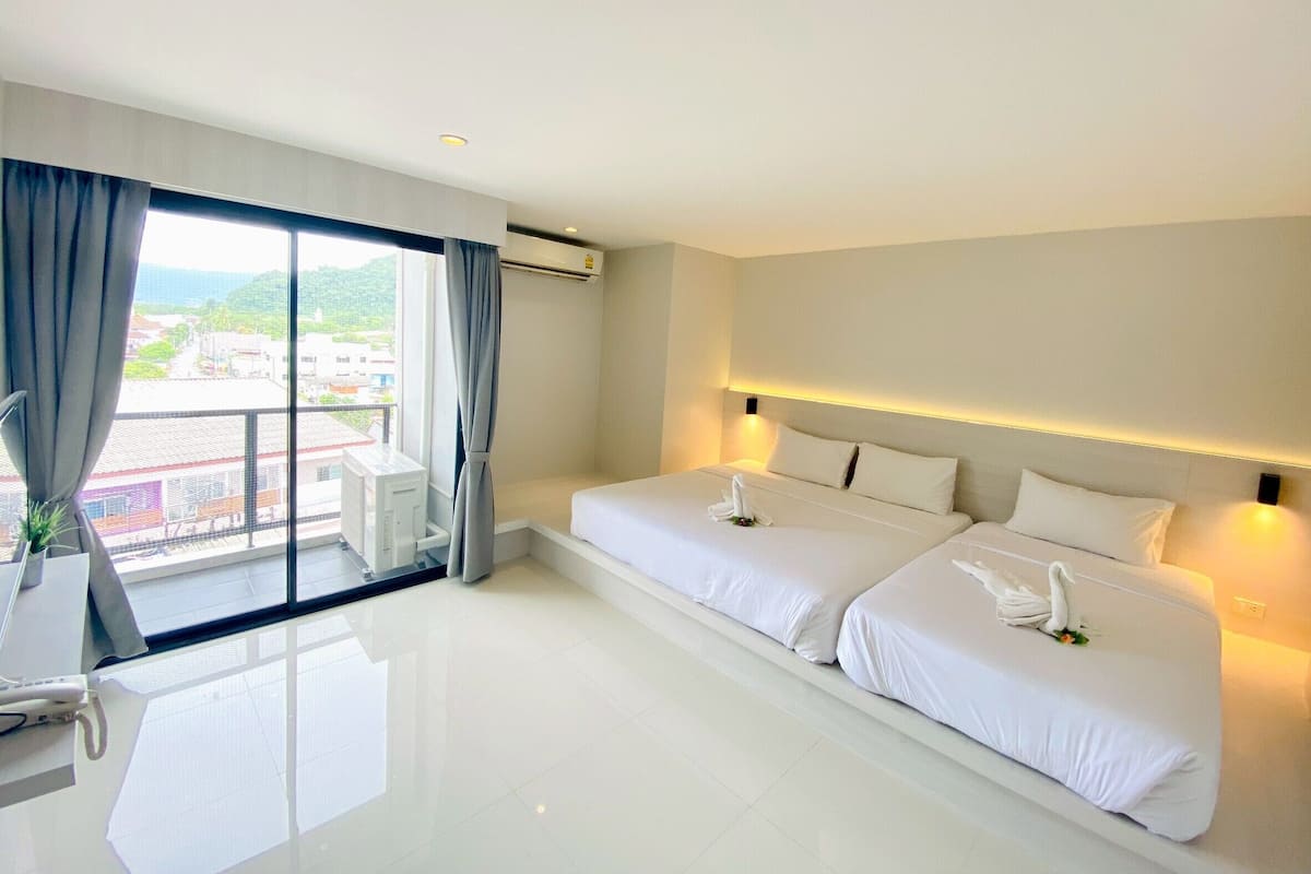 Quarto de hotel com duas camas, uma janela grande com cortinas, uma escrivaninha com cadeira e vista para a paisagem urbana.