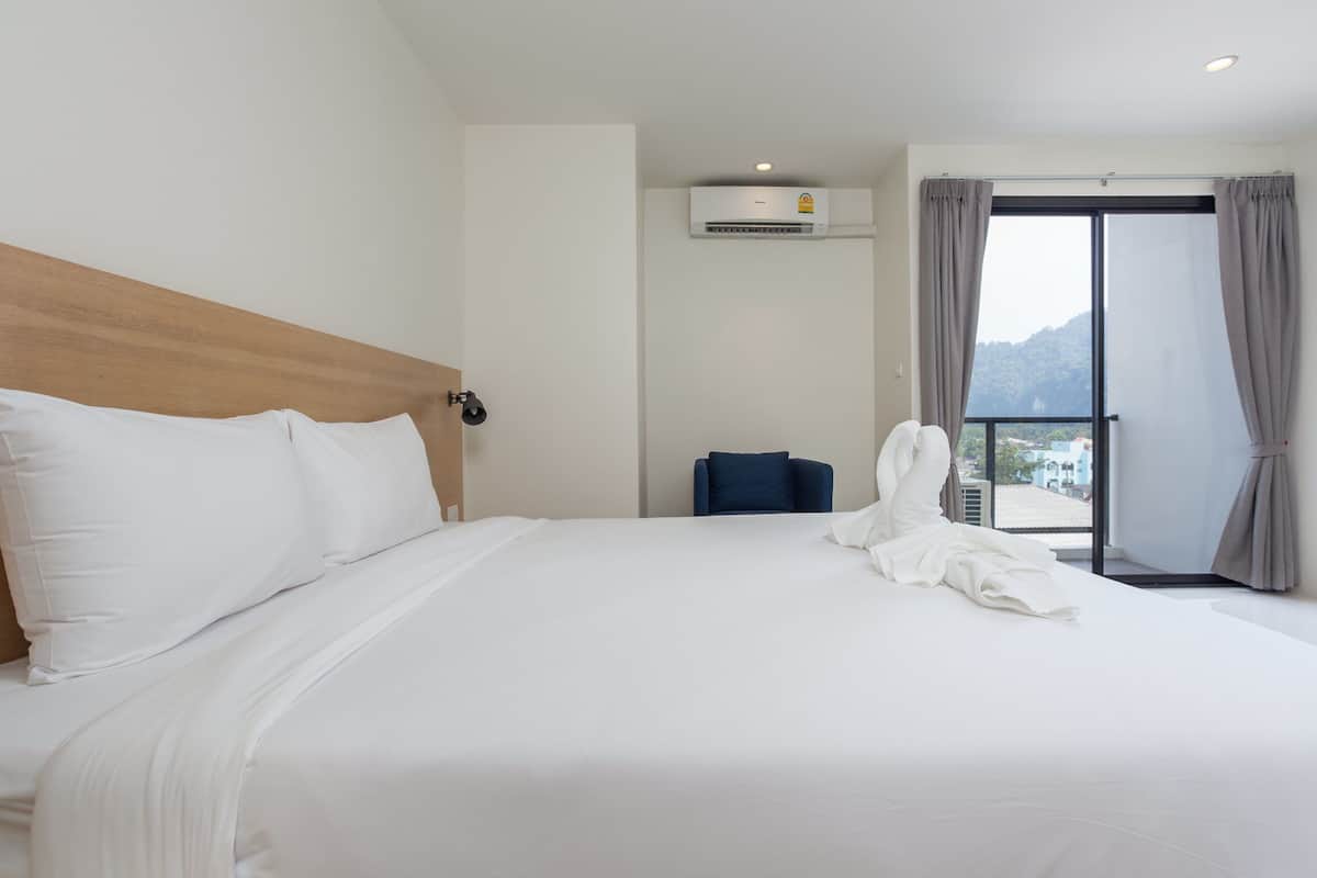Quarto de hotel com uma cama grande, um aparelho de ar condicionado, uma cadeira azul e vista para o exterior através de uma porta de correr.