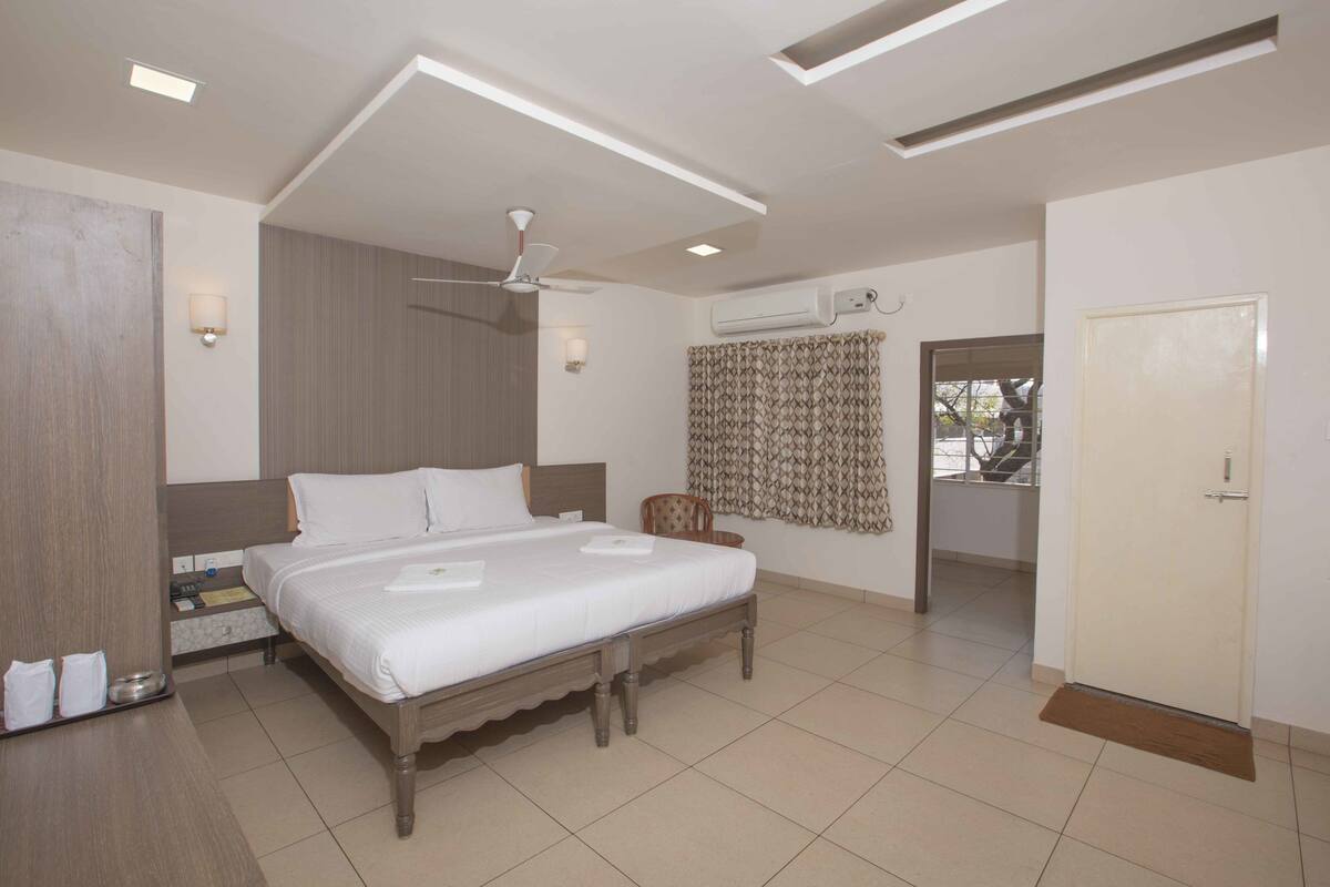 A hotel room with a large bed, a ceiling fan, an air conditioning unit, and a door leading to another room.