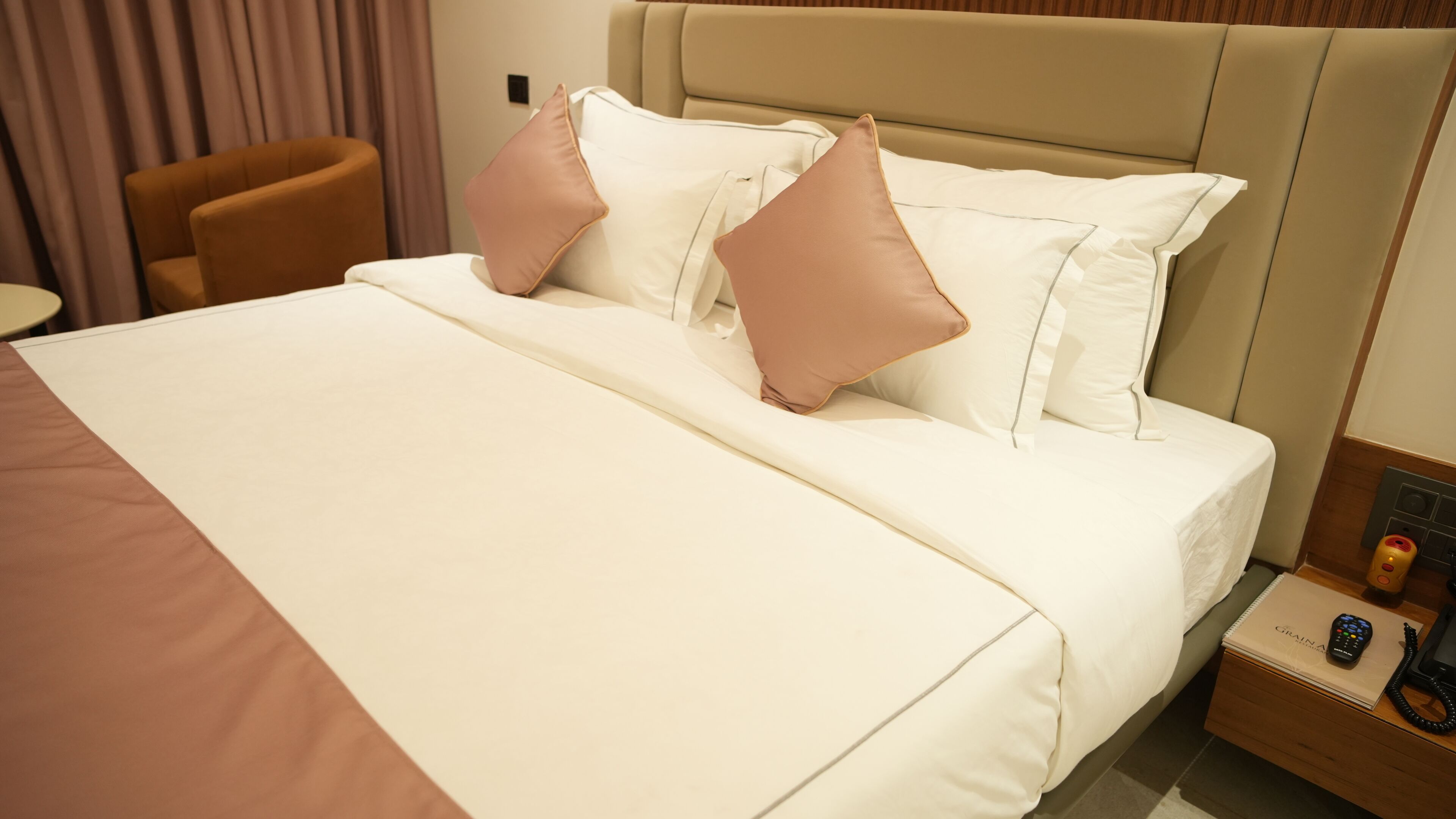 Habitación de hotel con una cama grande, dos almohadas, una mesita de noche con un teléfono y un control remoto, y una silla.