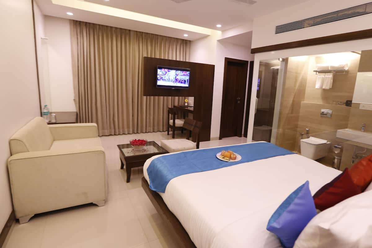 Ein Hotelzimmer mit Bett, Sofa, Fernseher, einem kleinen Tisch mit Obstschale und einem Badezimmer mit Waschbecken und Spiegel.