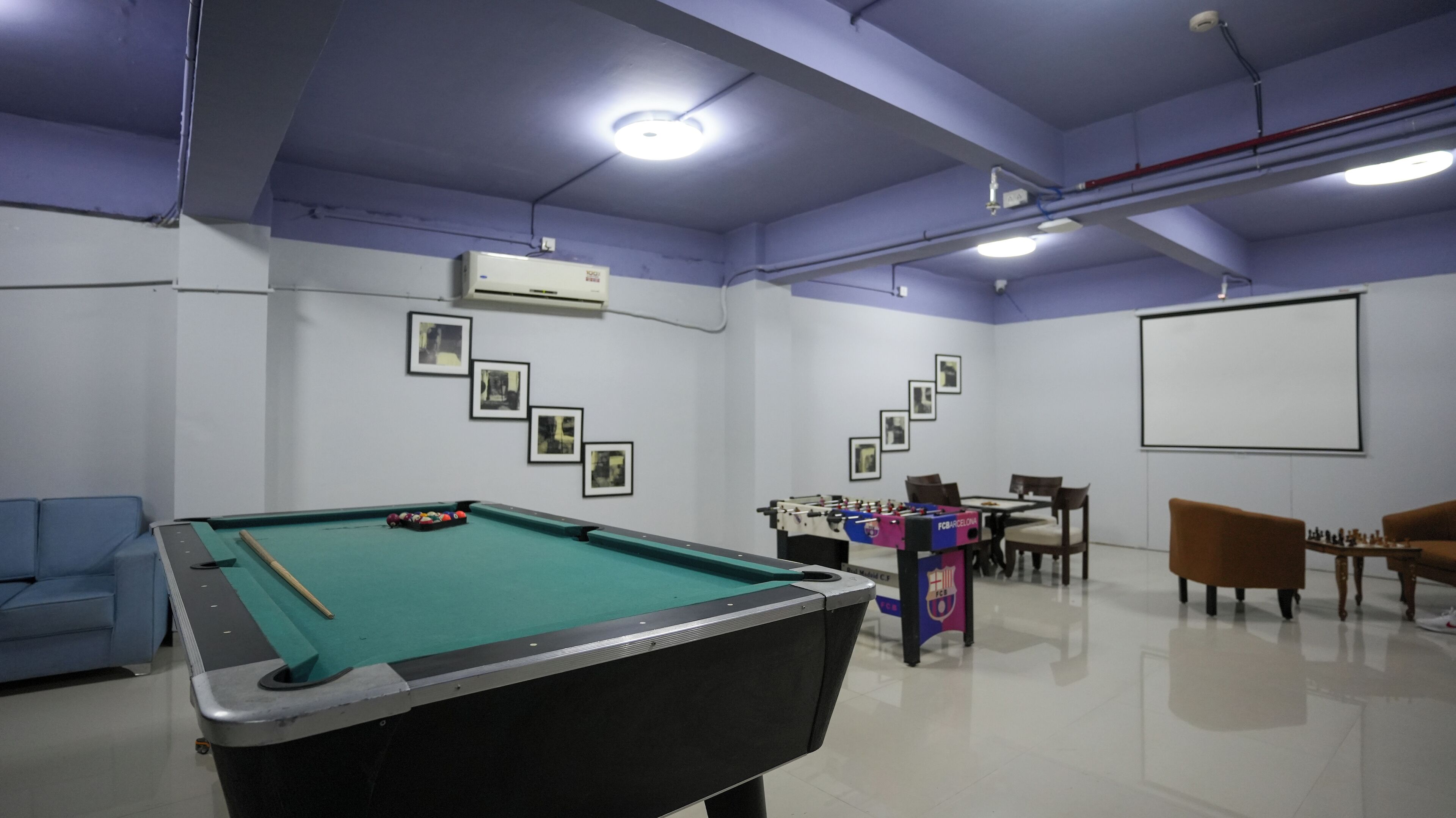 A pool table with a foosball table and chess set in a room with a projector screen and framed pictures on the wall.