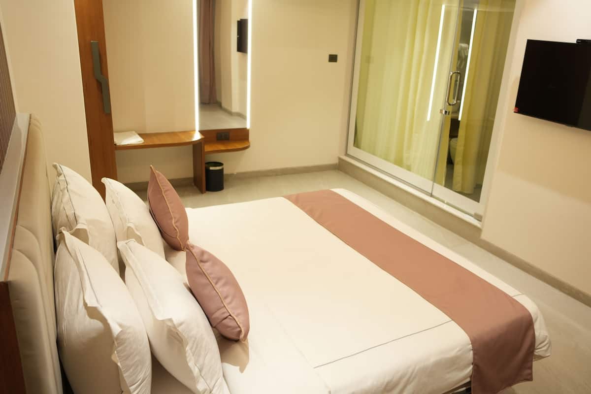 Ein Hotelzimmer mit Bett, Kissen, Nachttisch, Spiegel, an der Wand montiertem Fernseher und einem Fenster mit Vorhängen.