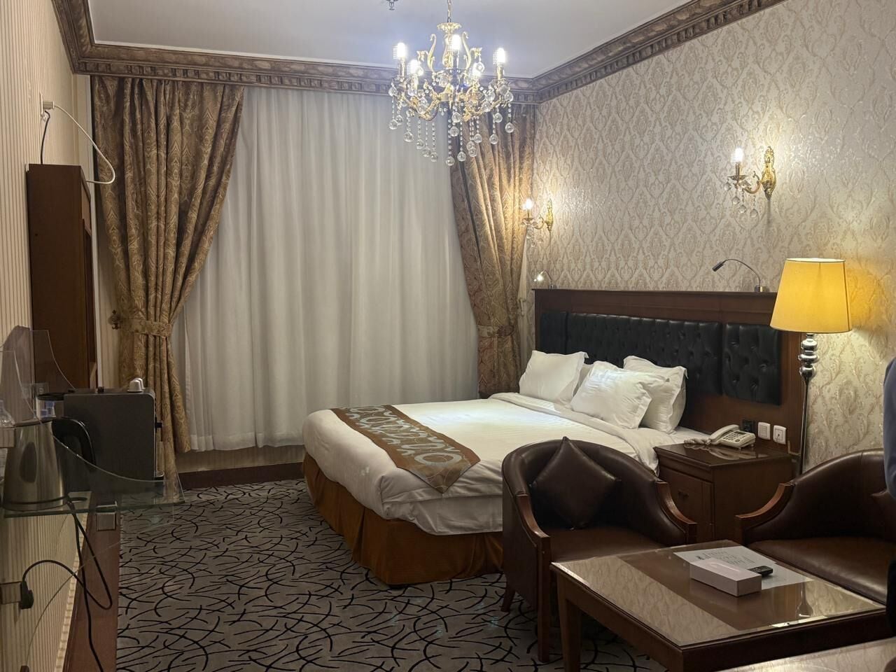 A hotel room with a large bed, a desk, a chair, a nightstand, a lamp, and a chandelier.