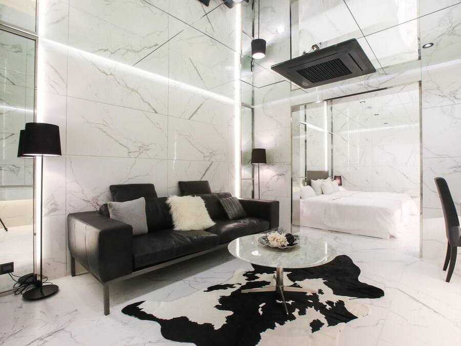 Una habitación de hotel moderna con sofá de cuero negro, una mesita de centro redonda, cama blanca y cabina de ducha de vidrio.