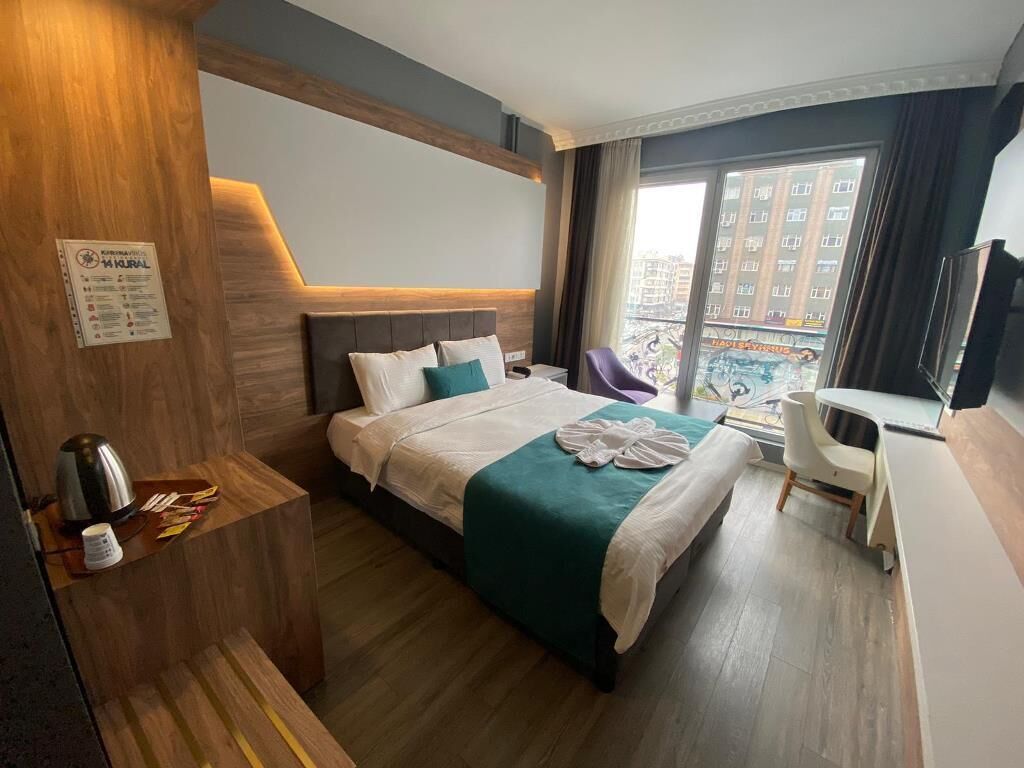 A hotel room with a bed, bedside table, and a view of the city through a large window.