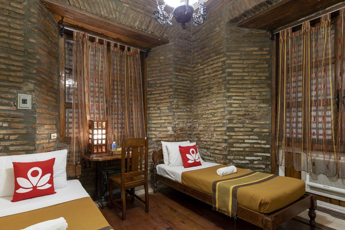 A room with two beds, a wooden chair, a small table, and a brick wall.