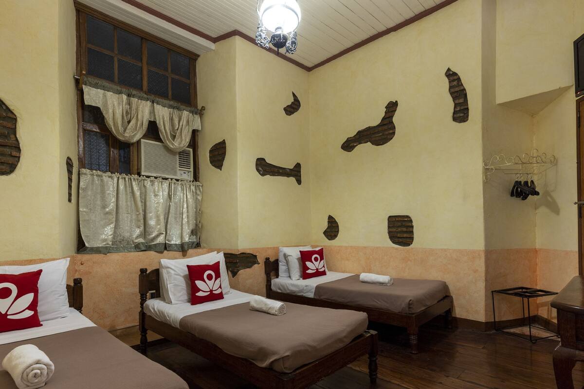 A room with two single beds, a window with curtains, a ceiling fan, and a brick wall decoration.