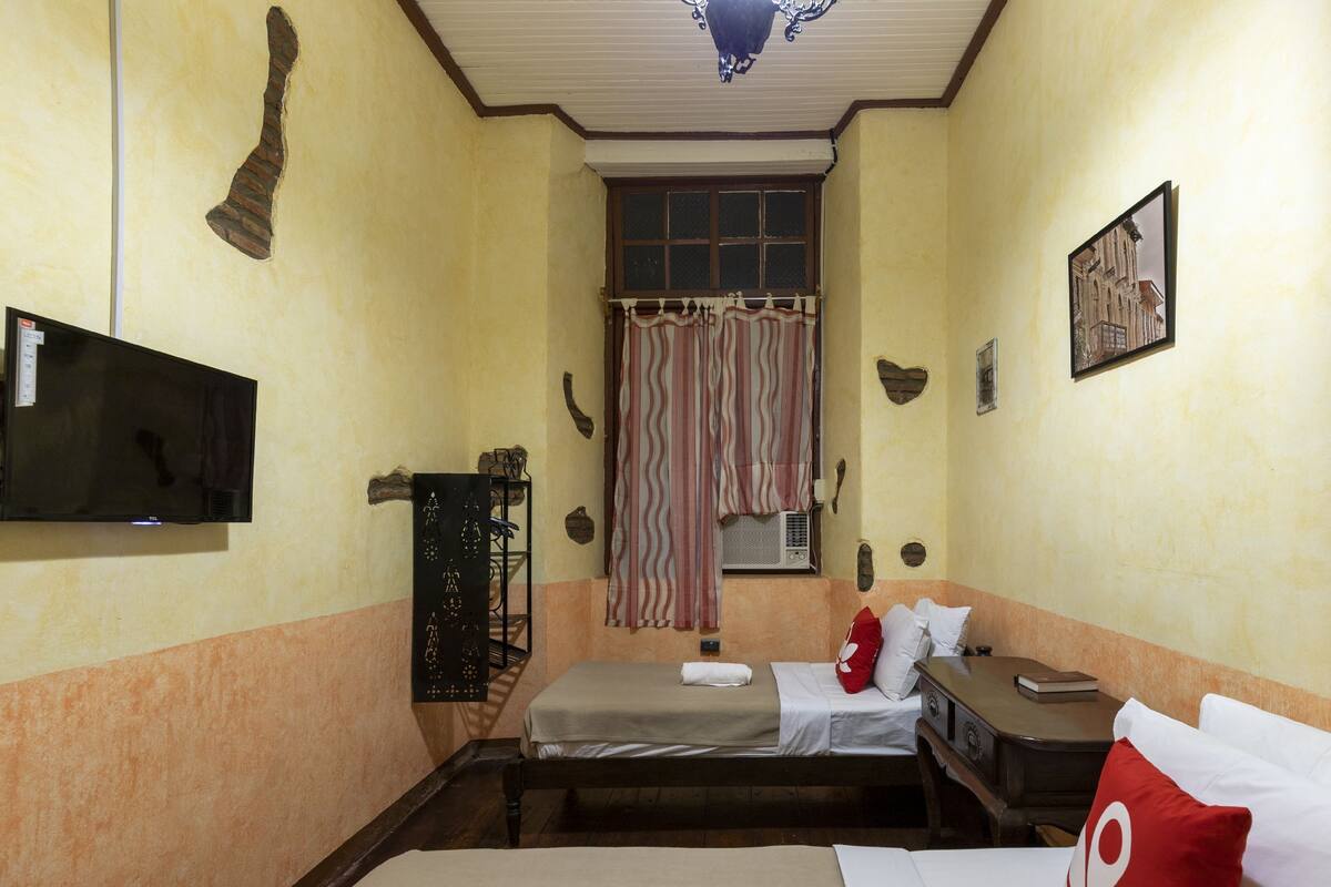 A small, compact room with a single bed, a desk, a chair, a television, and a window with curtains.