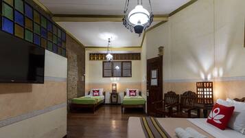 Habitación de hotel con dos camas individuales, una cama grande, televisor, armario de madera y ventana de estilo tradicional con celosía decorativa.