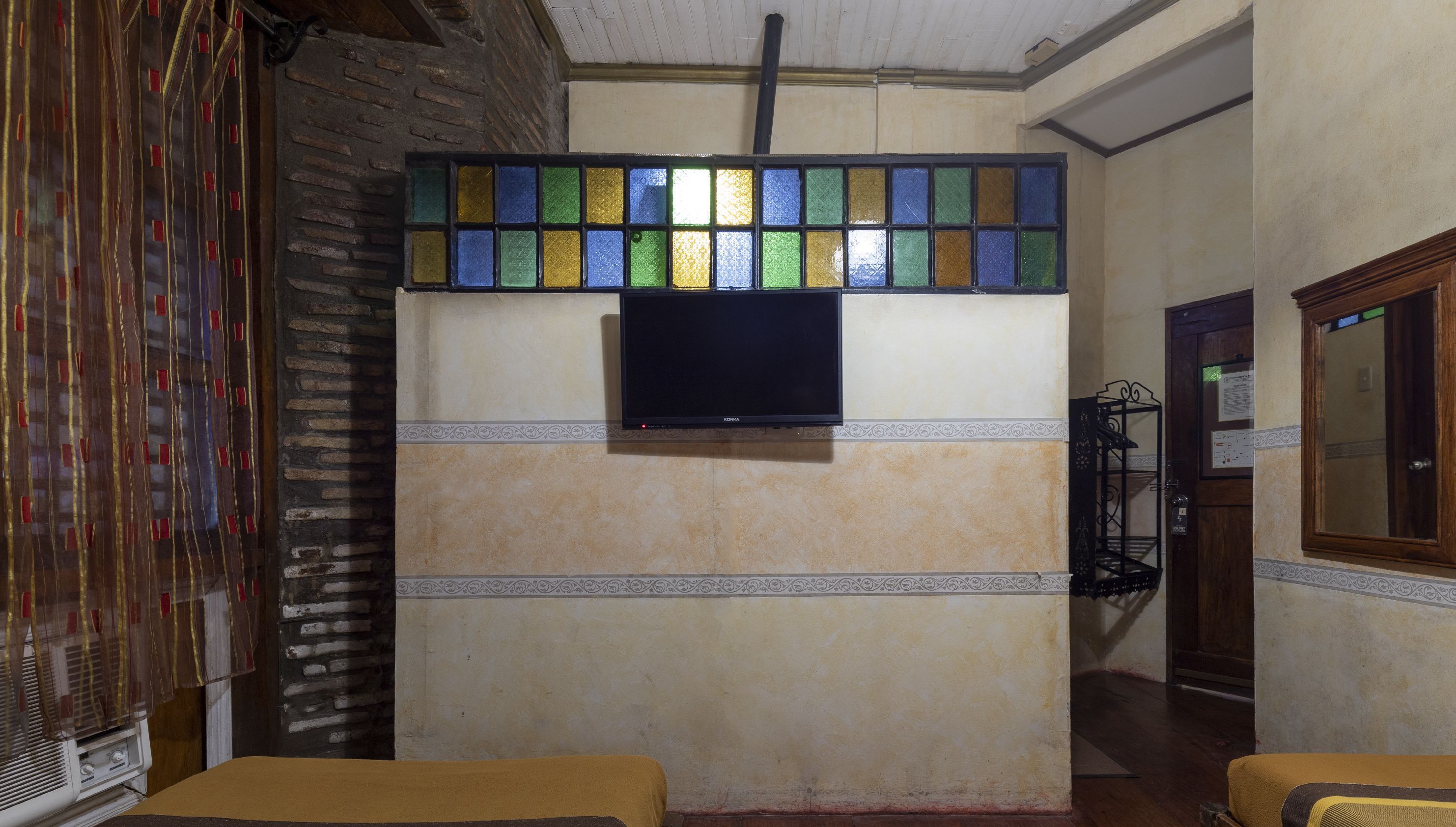 Una habitación con un televisor montado en la pared sobre un diseño de baldosas estampadas, una puerta de madera y un estante metálico con objetos a la derecha.