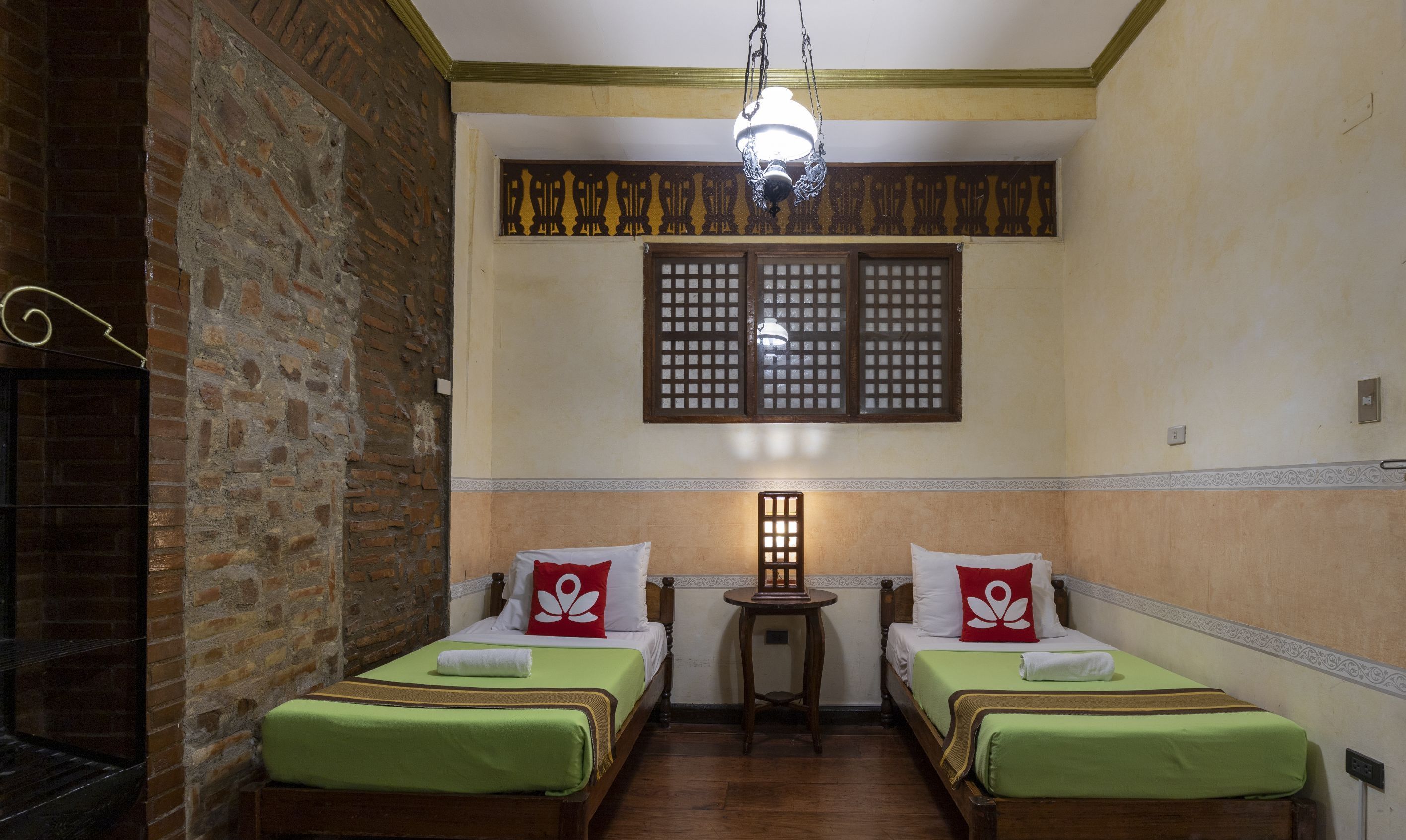 A room with two single beds, a small table, a decorative window, and a hanging light fixture.