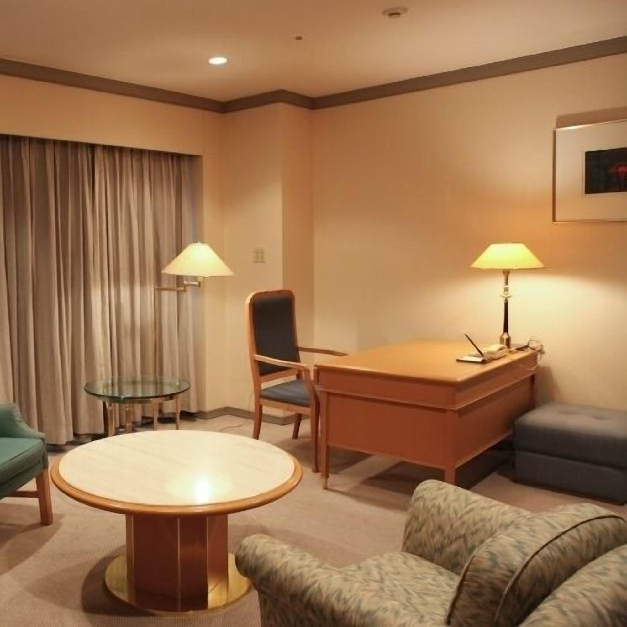 A hotel room with a round table, a desk, a chair, a sofa, a glass table, and two lamps.