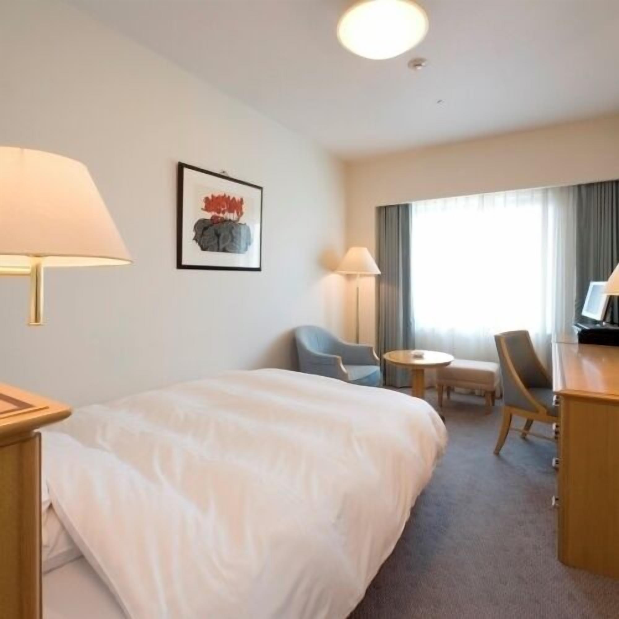 A hotel room with a bed, a desk, a chair, a small table, and a lamp.