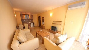 A living room with a sofa set, a coffee table, and a television. There is a kitchen area in the background with wooden cabinets and a countertop. - Menada Sea Regal Apartments (Sunny Beach)