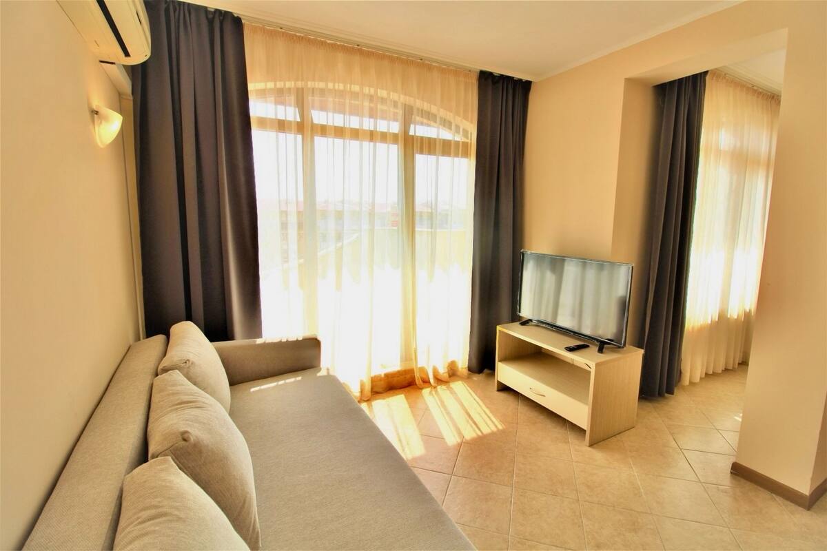 A modern hotel room with a sofa, a TV on a wooden stand, and large windows with sheer curtains.
