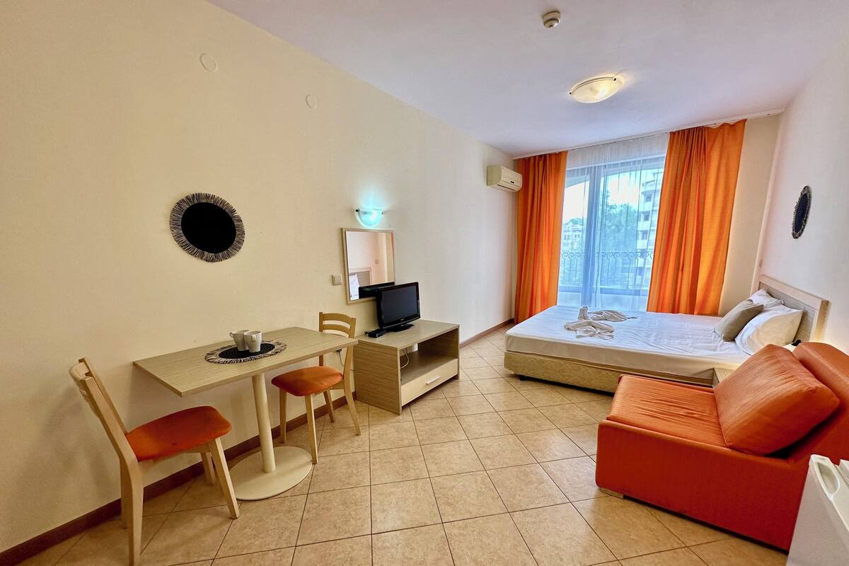 A hotel room with a bed, a desk with a computer, a chair, a small table with a cup, and a red sofa.