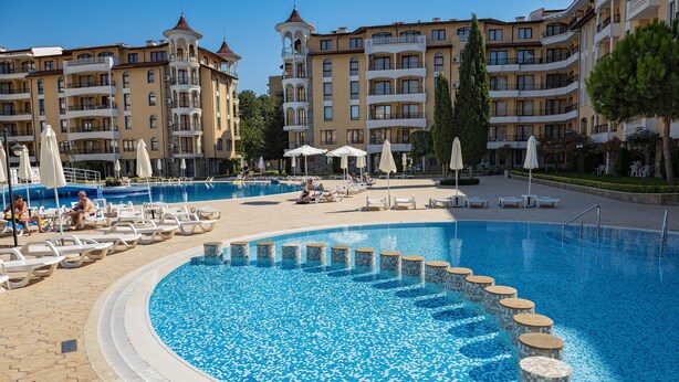 A pool area with lounge chairs and umbrellas, surrounded by multi-story buildings with balconies. - Menada Royal Sun Apartments (Sunny Beach)