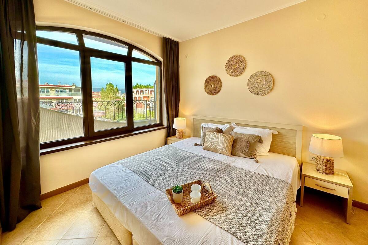 A hotel room with a large bed, two bedside tables, a window with curtains, and decorative wall art.