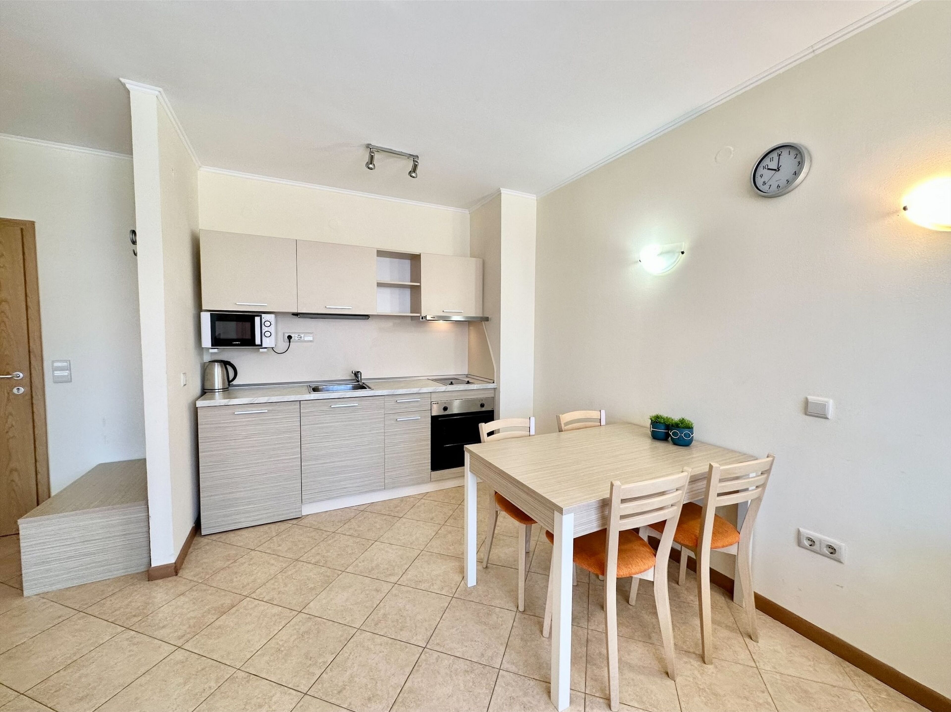 A compact kitchen with a dining area, a microwave, a kettle, and a clock on the wall.