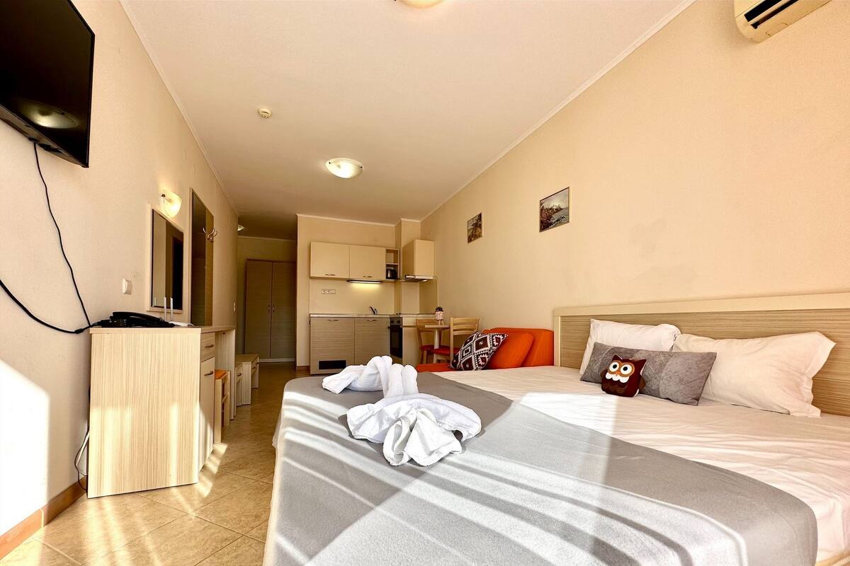 A hotel room with a bed, a desk, a kitchenette, and a TV mounted on the wall.