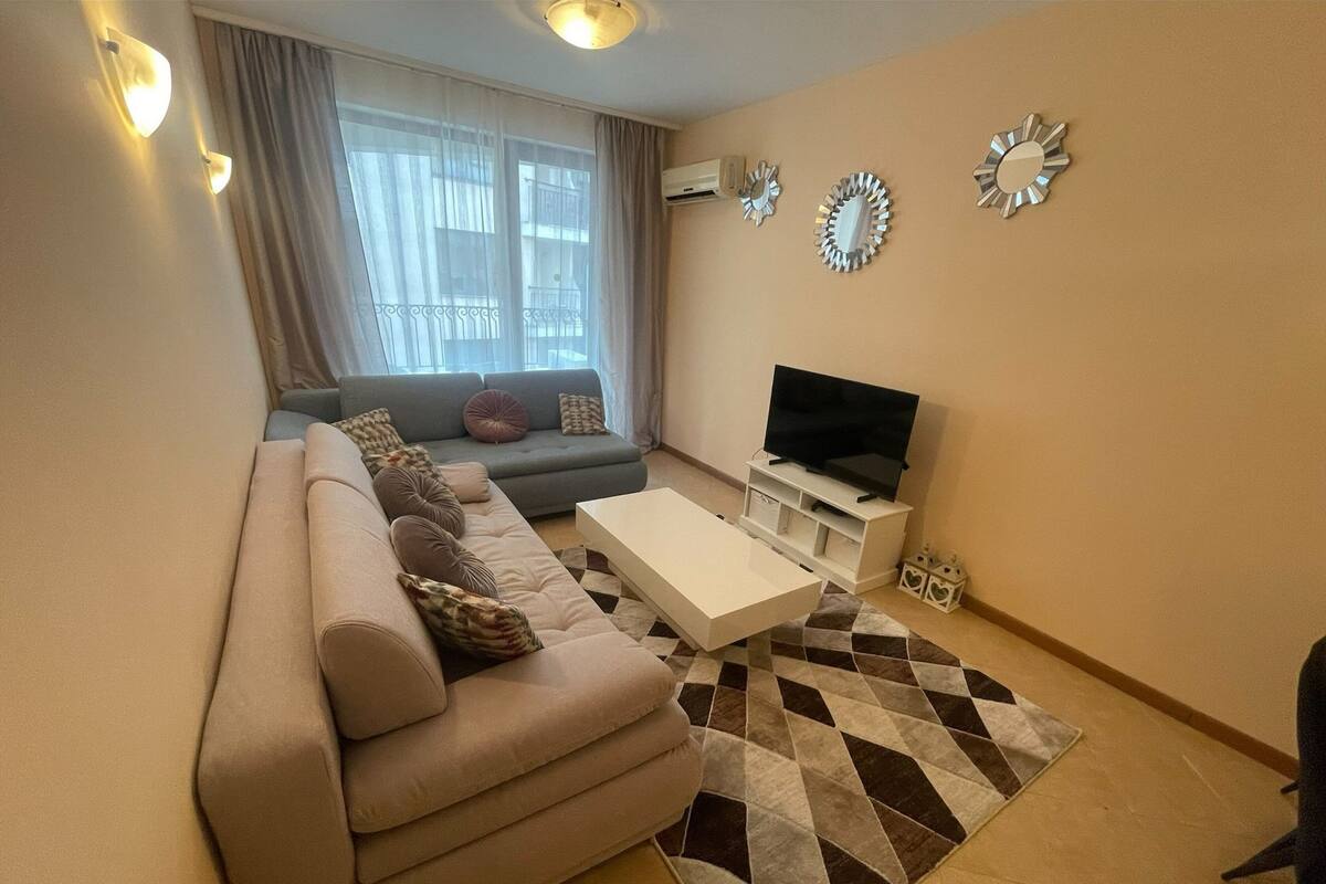 A living room with a sofa, a coffee table, a TV on a stand, decorative mirrors, and a window with curtains.