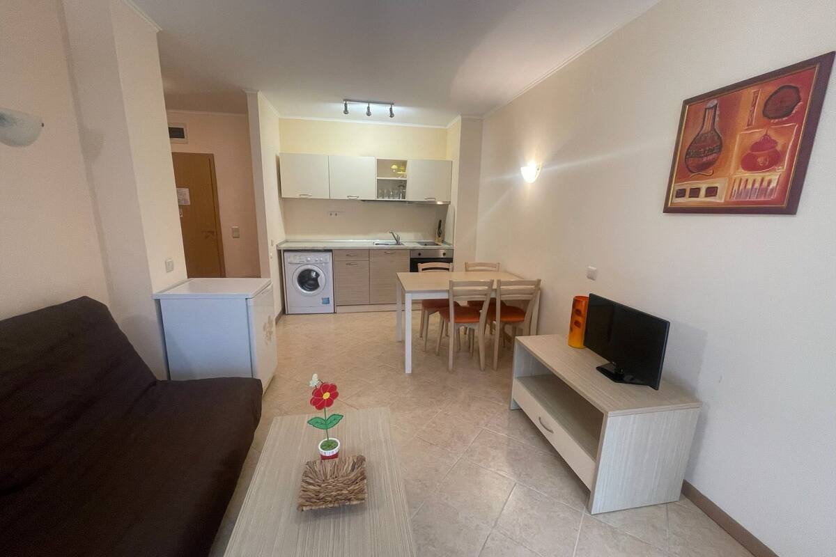 A compact living space with a sofa, a TV stand, a dining table with chairs, a washing machine, and a kitchen area.