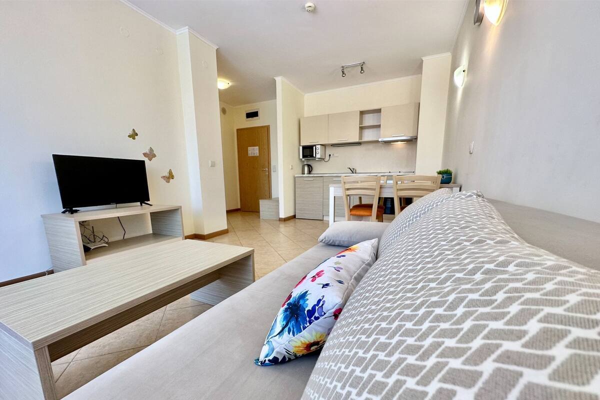 A modern living room with a sofa, a TV on a stand, and a kitchen in the background.