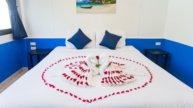 Habitación de hotel con una cama decorada con un arreglo de toallas con forma de corazón y pétalos de rosa, un cuadro de una playa en la pared, y una decoración en tonos azul y blanco.