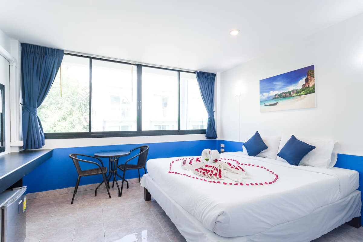 A hotel room with a large bed, a small table, and a chair. There is a window with curtains, a painting on the wall, and a view of a beach.