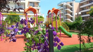 A playground with a slide and climbing structures, surrounded by buildings and greenery. - Menada Grand Kamelia Apartments (Sunny Beach)