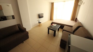 A living room with a sofa, a coffee table, a TV, and a window with curtains. - Menada Global Ville Apartments (Sunny Beach)