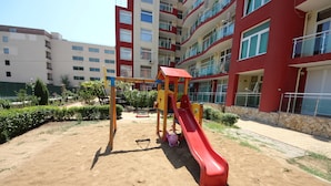 A playground with a red slide and yellow structures, surrounded by a sandy area with greenery and a multi-story building in the background. - Menada Global Ville Apartments (Sunny Beach)