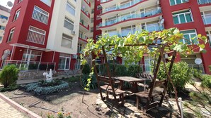 A multi-story residential building with balconies, a garden area with a wooden pergola, and a statue. - Menada Global Ville Apartments (Sunny Beach)
