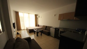 A compact living space with a sofa, a small dining table, a kitchenette, and a large window with curtains. - Menada Global Ville Apartments (Sunny Beach)