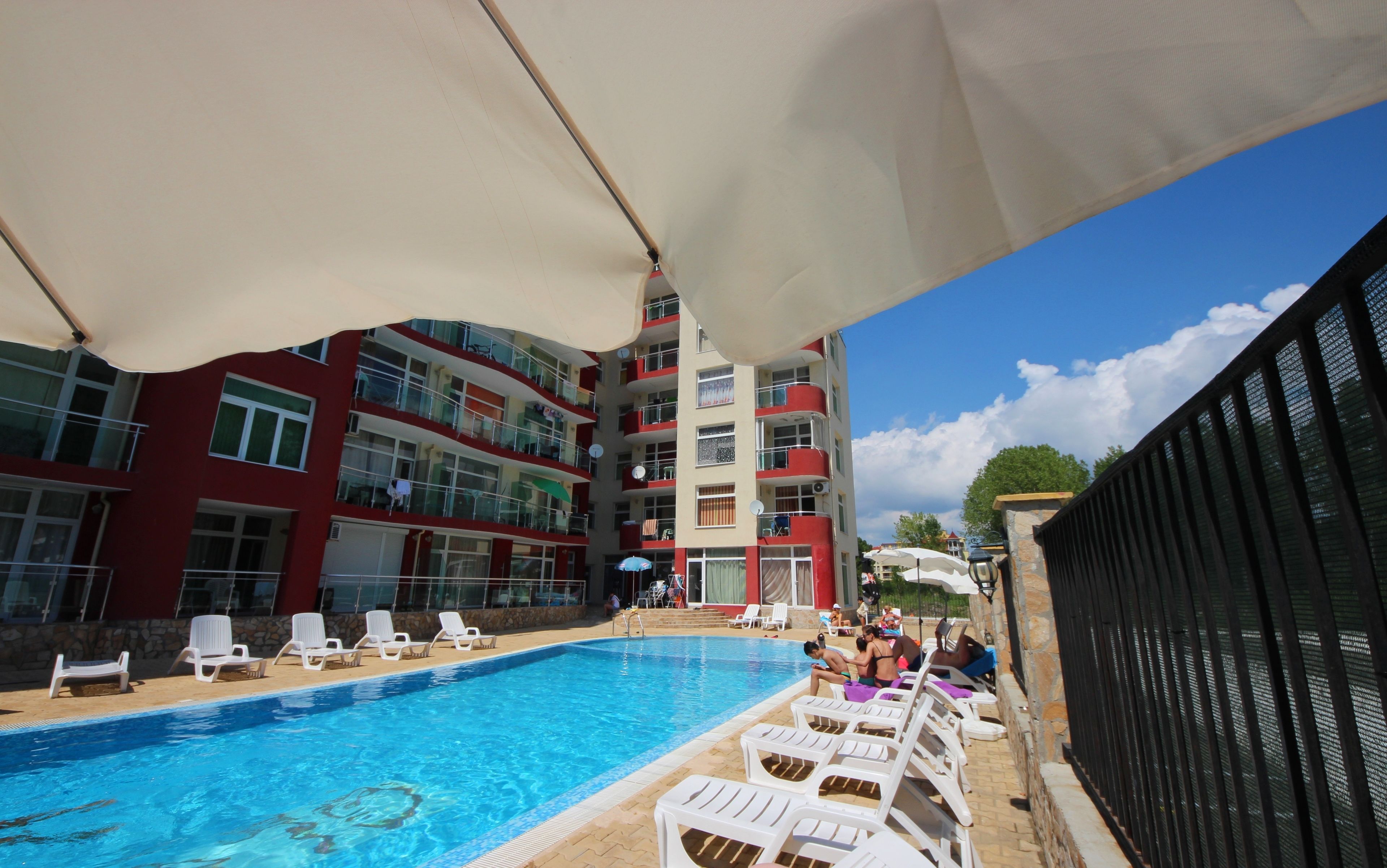 A swimming pool with white lounge chairs, a red and beige building with balconies, and a clear blue sky.