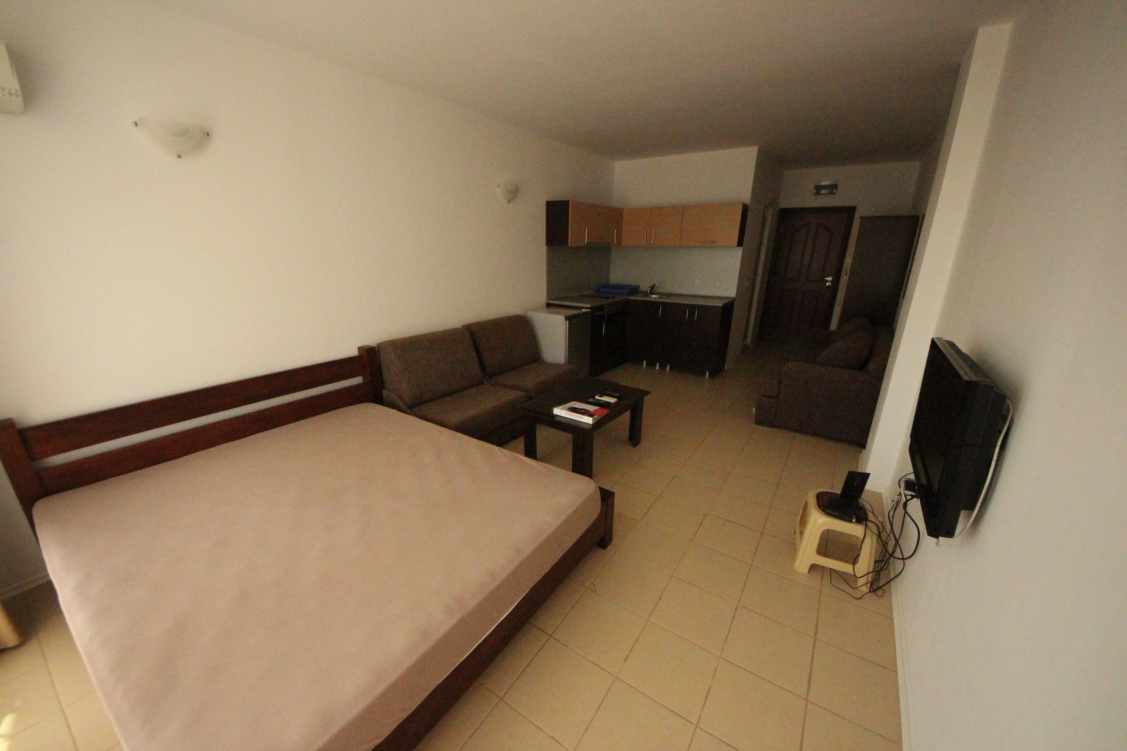 A bedroom with a bed, a sofa, a small table, a TV mounted on the wall, and a kitchenette in the background.