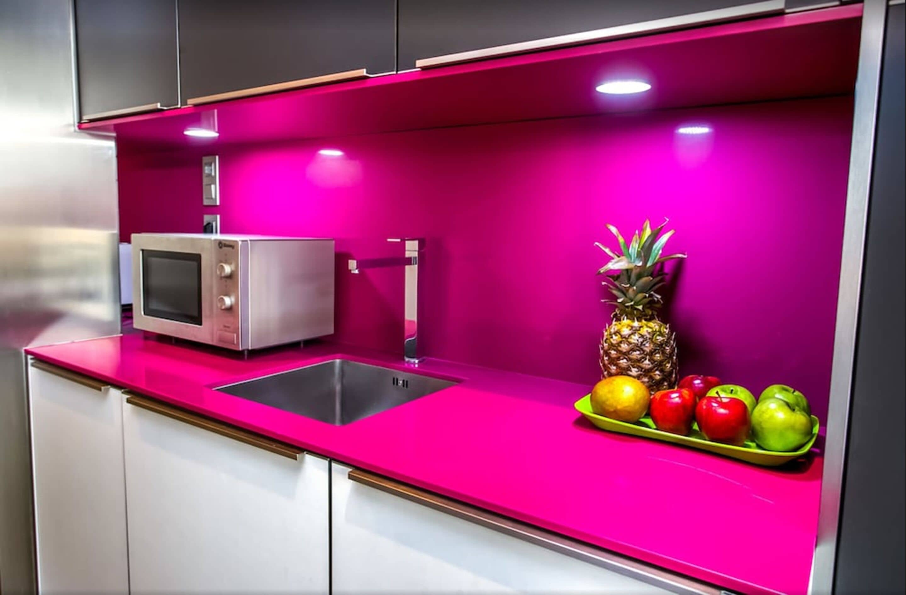 A kitchen with a pink countertop, a microwave, and a fruit bowl with apples and a pineapple.