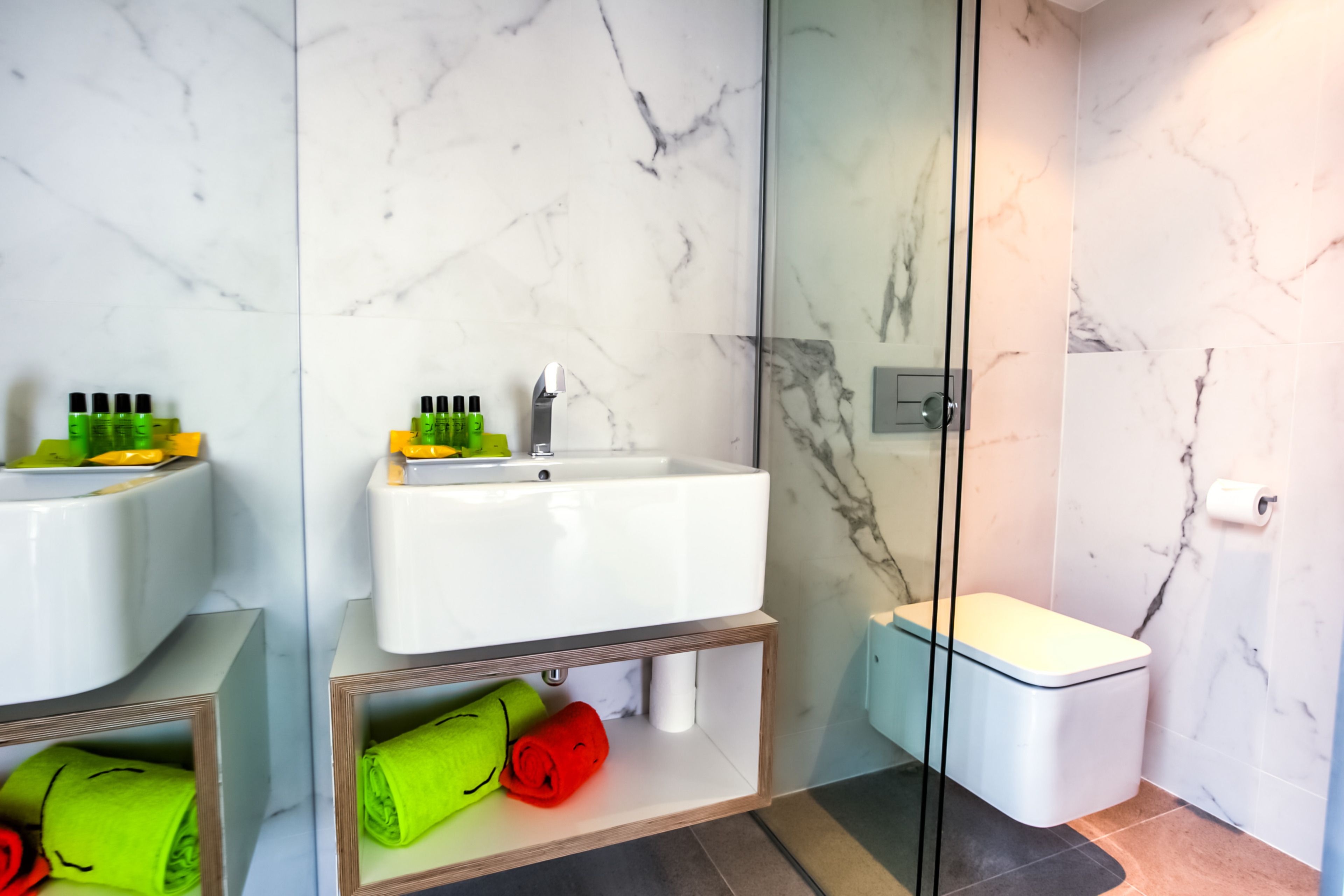 A modern bathroom with a white sink, mirror, and toilet. There are green and red towels on the shelves.