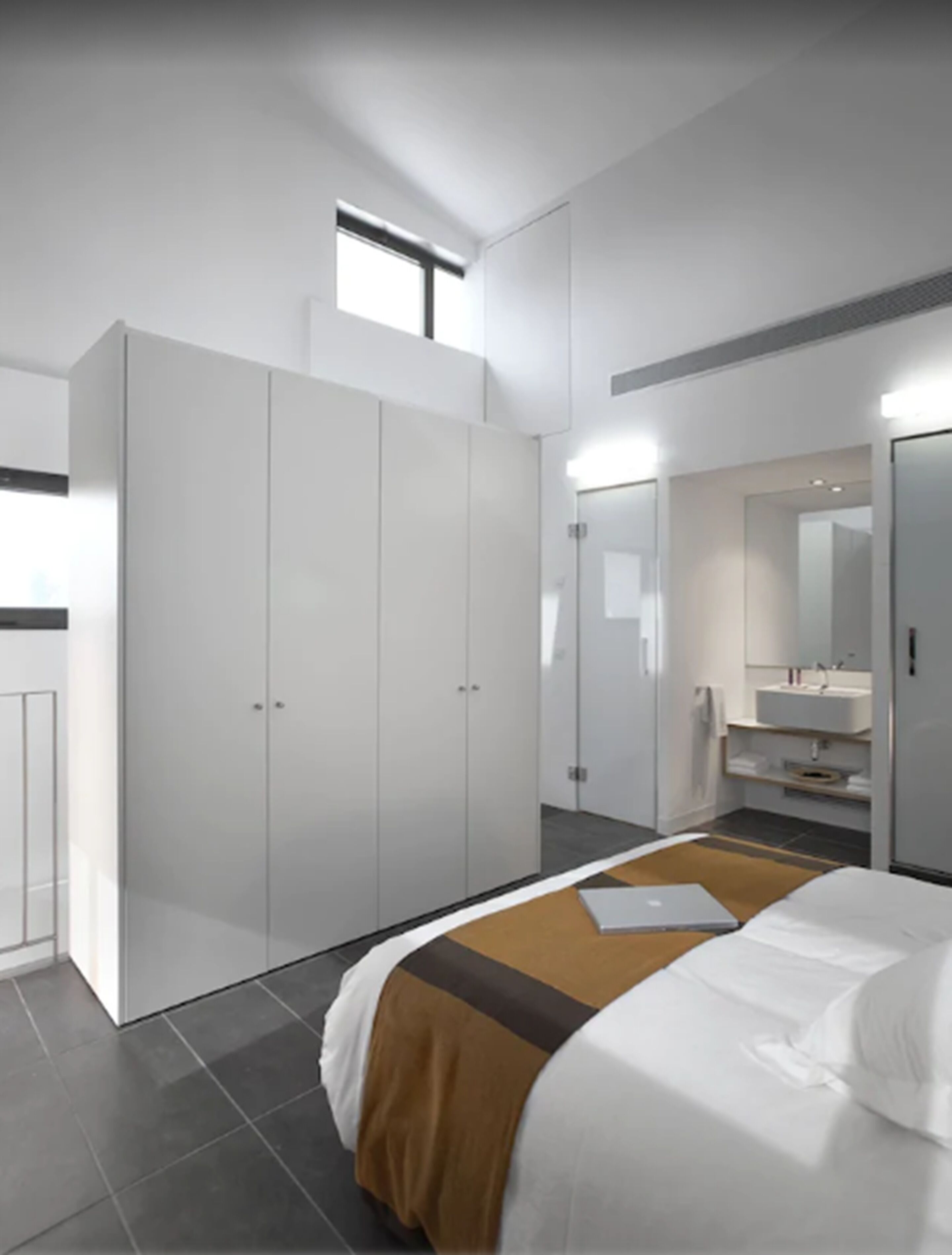 Un dormitorio moderno con un armario grande, una cama con colcha marrón y blanca, un baño con espejo y lavamanos, y un ventanal pequeño.